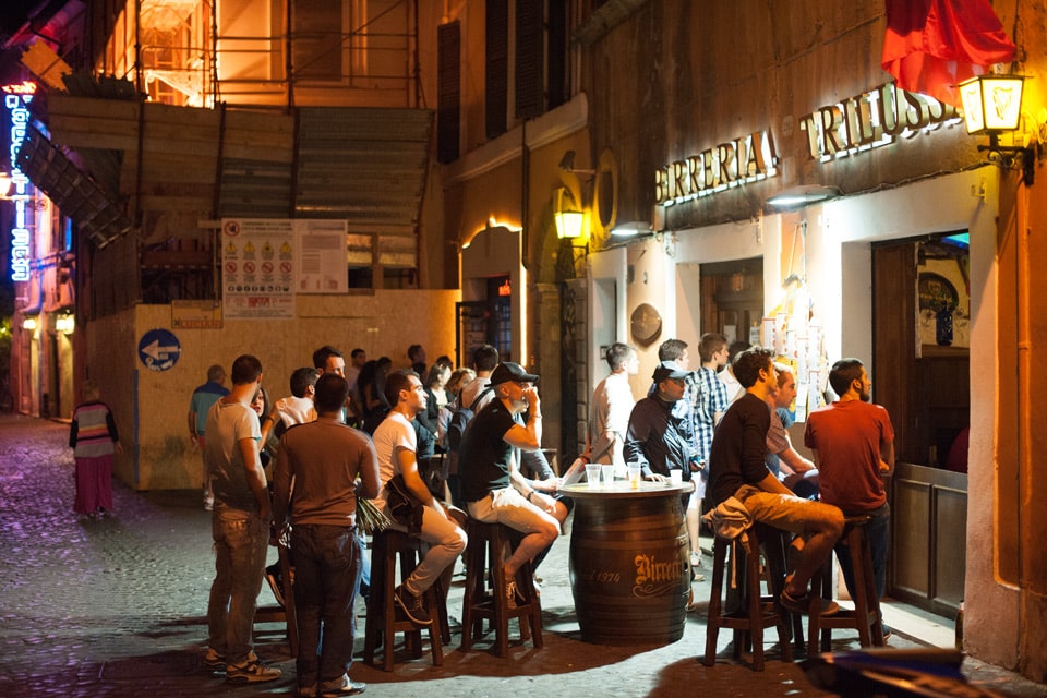 Personas alrededor de mesas hechas con barriles frente a la cervecería Birreria Trilussa por la noche, Trastevere, Roma